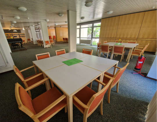 Ansicht der Gemeinschaftsräume im Foyer. Mehrere Tischgruppen, Bücherregale und ein Klavier laden zu verweilen ein. Der Raum bietet eine schöne Aussicht nach außen.